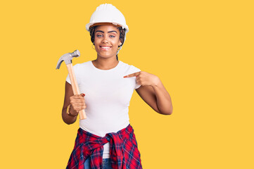 Young african american woman with braids wearing hardhat holding hammer pointing finger to one self...