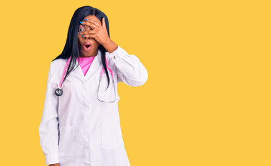 Young african american woman wearing doctor stethoscope peeking in shock covering face and eyes with hand, looking through fingers with embarrassed expression.