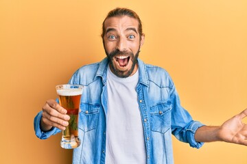Attractive man with long hair and beard drinking a pint of beer celebrating victory with happy smile and winner expression with raised hands