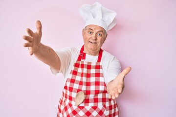 Senior grey-haired man wearing professional baker apron looking at the camera smiling with open arms for hug. cheerful expression embracing happiness.