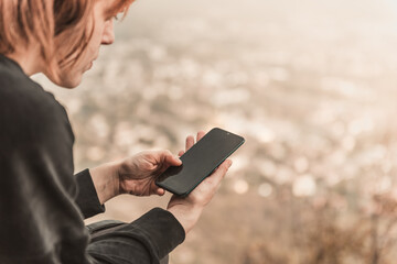 The red-haired traveler girl writes SMS on her smartphone sitting on the mountain rock. The concept of travel, technology.