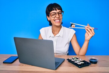 Beautiful brunettte woman working at the office eating sushi winking looking at the camera with...