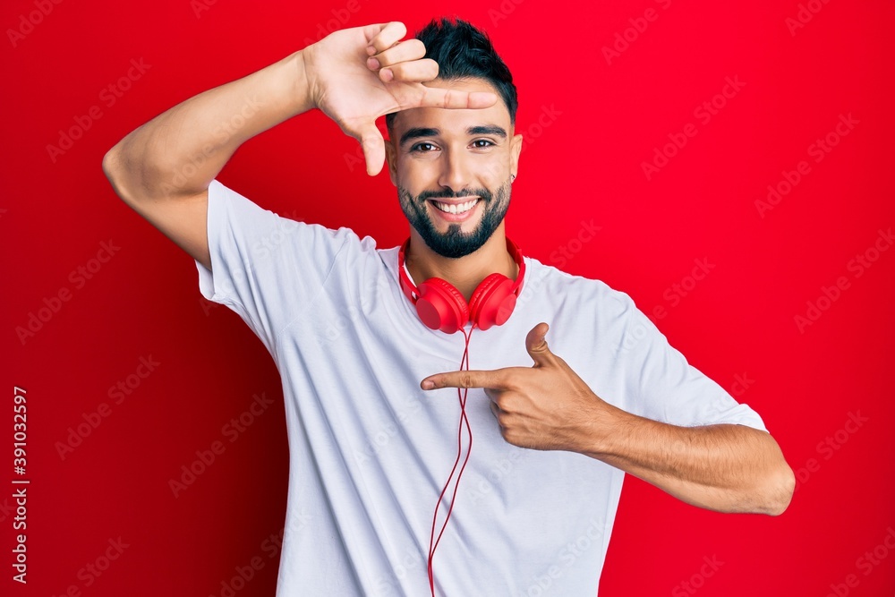 Wall mural Young man with beard listening to music using headphones smiling making frame with hands and fingers with happy face. creativity and photography concept.