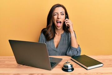 Young brunette woman working at hotel reception talking on the phone winking looking at the camera...