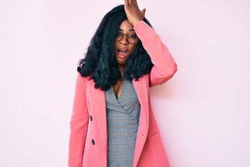 Beautiful african woman wearing business jacket and glasses surprised with hand on head for...