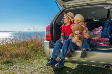 Family vacation by the sea. Two boys are sitting in the trunk of a car. The tailgate is open up. The youngest of the brothers is holding a teddy bear toy.