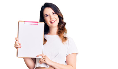 Beautiful young brunette woman holding clipboard with blank space looking positive and happy standing and smiling with a confident smile showing teeth