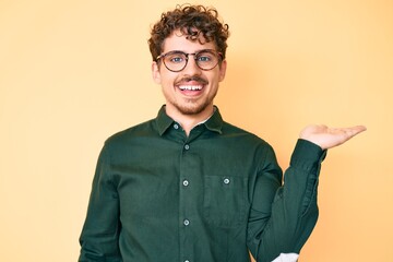 Young caucasian man with curly hair wearing casual clothes and glasses smiling cheerful presenting and pointing with palm of hand looking at the camera.