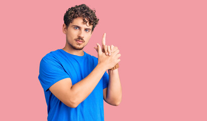 Young handsome man with curly hair wearing casual clothes holding symbolic gun with hand gesture, playing killing shooting weapons, angry face