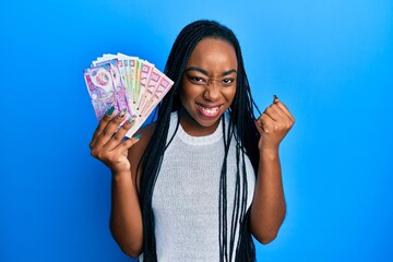 Young african american woman holding hong kong dollars banknotes screaming proud, celebrating victory and success very excited with raised arm