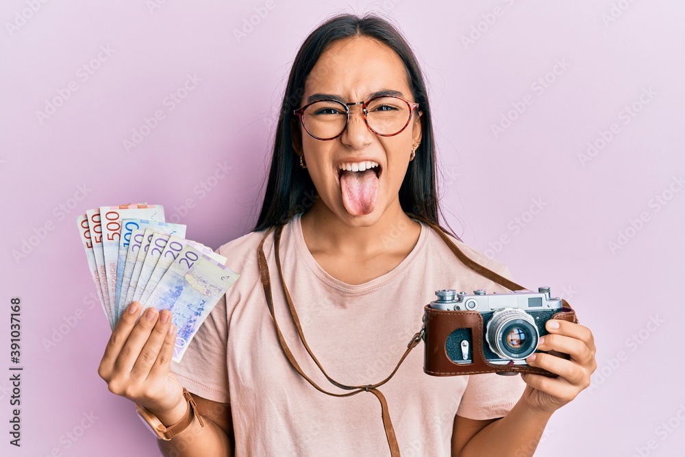 Sticker Young asian woman holding vintage camera and swedish krona sticking tongue out happy with funny expression.