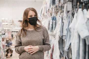 Young beautiful pregnant woman in hygienic mask choosing baby clothes, stroller or pram buggy for newborn. Shopping for expectant mothers and baby. Preventive measures on pandemic lifestyle.