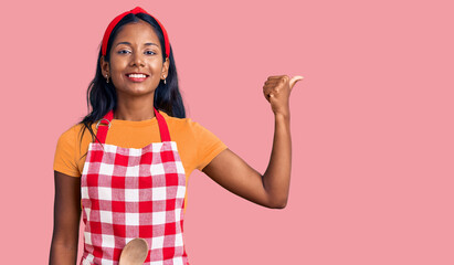 Young indian girl wearing professional baker apron smiling with happy face looking and pointing to the side with thumb up.