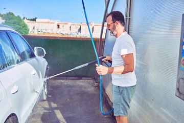 Middle age handsome man wearing casual clothes and sunglasses smiling happy. Standing with smile on face washing car using water pistol.