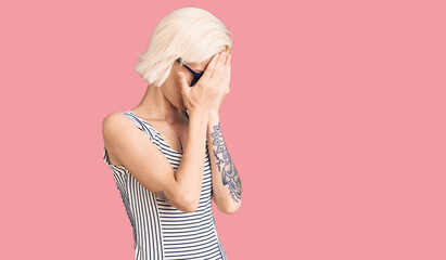 Young blonde woman with tattoo wearing casual clothes and glasses with sad expression covering face with hands while crying. depression concept.