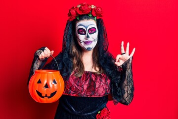 Young woman wearing day of the dead costume holding pumpkin doing ok sign with fingers, smiling friendly gesturing excellent symbol