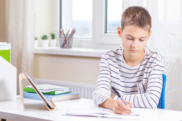 Kid with digital tablet computer writing, doing homework at white desk. Online learning, remote education, distance lessons at home