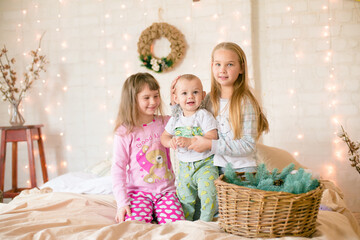 Cute sisters and little brother  in pajamas have fun in in a Scandinavian style bedroom decorated with Christmas garlands and needles on a large bright bed. Christmas mood. Christmas mood. Family time