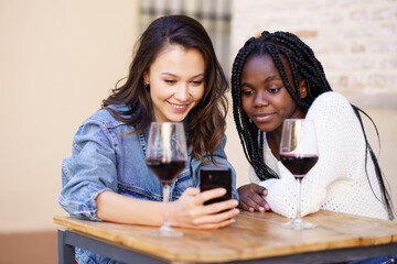 Two women looking at their smartphone together while having a glass of wine.