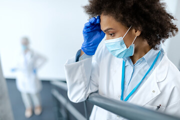 Distraught African American female doctor with protective face mask at hospital hallway.