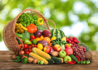 Overturned wicker basket with different fresh organic vegetables and fruits on wooden table against...
