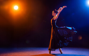 Ballerina in a black dress and crown is dancing on black background illuminated by multicolored beams of spotlights