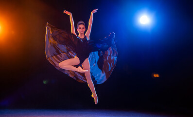 Ballerina in a black dress and a crown flying in a jump on black background illuminated by multicolored beams of spotlights