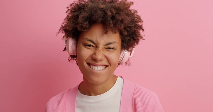 Joyous dark skinned woman smiles broadly sends positive vibes listens music in stereo headphones moves carefree poses against pink background enjoys great vibes isolated over pink background
