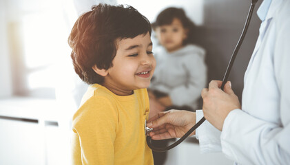 Woman-doctor examining a child patient by stethoscope in sunny clinik. Cute arab boy at physician appointment