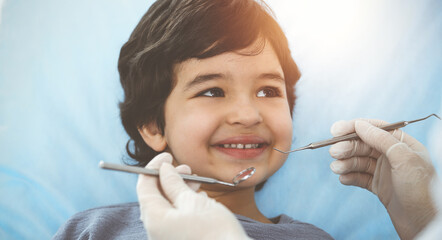 Cute arab boy sitting at dental chair with open mouth during oral checking up with doctor. Visiting sunny dentist office