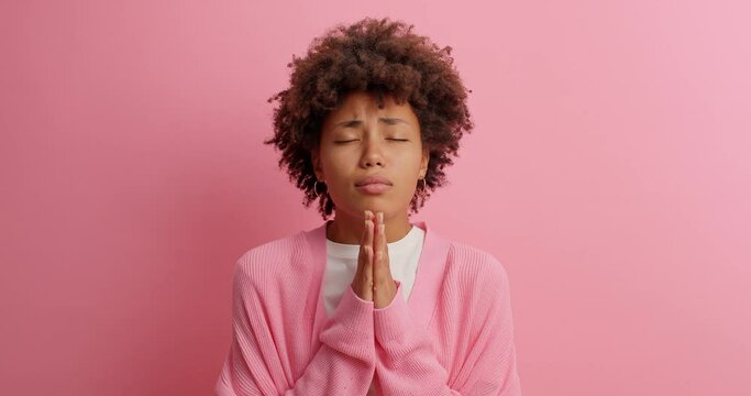 Sad dark skinned young woman keeps palms in pray gesture looks with begging expression asks for offer has imploring look dressed casually isolated over rosy background. Please one more favor
