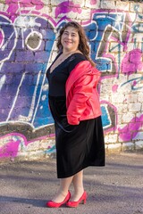 Sehr mollige Frau mittleren Alters in schwarzem Samtkleid und roter Lederjacke vor Mauer mit...