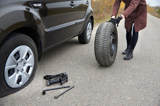 A young woman rolls spare tire near her car with a flat tire.