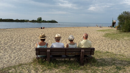 Gruppe von älteren Menschen sitzt auf einer Bank am Strand und blickt aufs Wasser