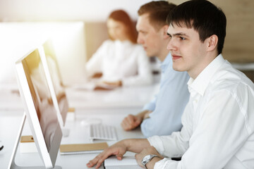 Cheerful smiling businessman at work in sunny office. Casual dressed entrepreneur using pc computer while sitting with diverse colleagues at the background