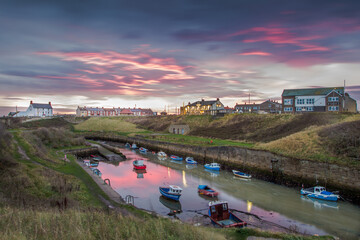 The Burn with the moored fishing boats of Seaton Sluice, Northumberland at sunrise, with the fiery sky reflecting in the water
