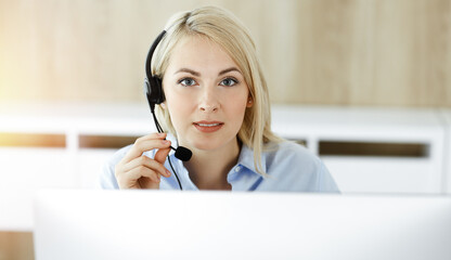 Blonde business woman sitting and communicated by headset in call center in sunny office. Concept of telesales business