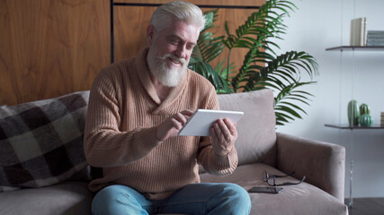 Reading News. Senior man with a gray beard using digital tablet sitting on the couch at home
