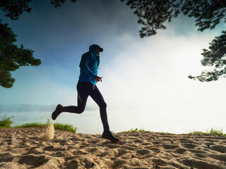 Male runner run hard alone in white sand of beach. Man wear blue sweatshirt and long black running leggings. Run along misty lake.