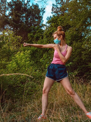 A girl in a mask works out a blow while standing in the middle of the forest. Sport in nature during the pandemic