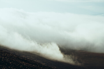 View from stony mountainside to highland valley within clouds. Atmospheric darker minimalist alpine landscape with low clouds in mountain valley. Wonderful mountain scenery with thick clouds on ground