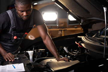 professional car mechanic is examining engine under the hood at auto repair shop, make notes,...