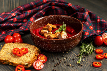 Thick tomato soup with meat slices in a ceramic plate. Next to the plate are a checkered serving napkin, bread, tomatoes, and a sprig of rosemary.