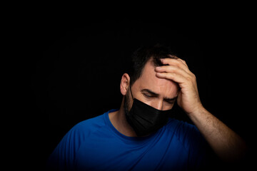 Bearded man dressed in blue t-shirt and with a black mask to protect himself from coronavirus posing against black background