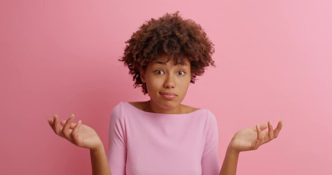 Bewildered hesitant young African American woman looks with confused puzzled face expression has doubtful expression shrugs shoulders faces difficult choice isolated over pink studio background.