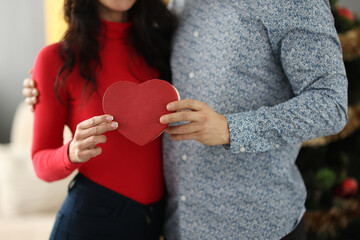 Woman and man are holding box with gift in form of heart in hands near Christmas tree close-up. Gifts for loved ones for New Year concept