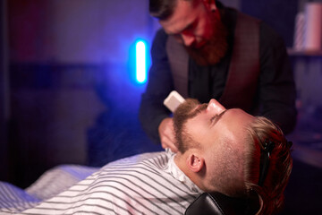 hairstyling process. barber cutting beard of caucasian male with the use of scissors, client sits in an armchair