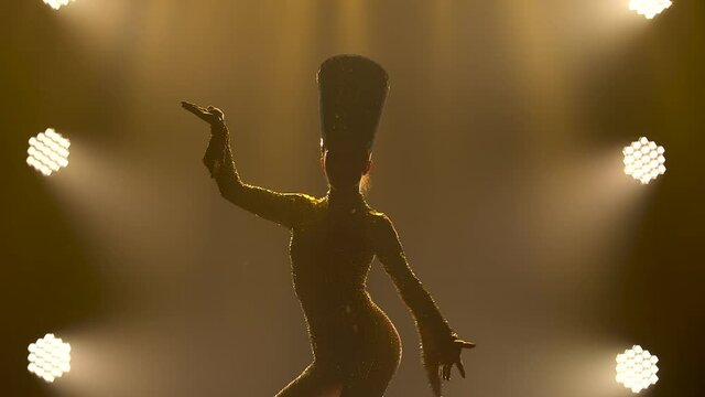 Silhouette of a young woman in a costume and a headdress of Cleopatra or Nefertiti dancing in a dark studio. Theatrical performance on a smoky background with studio lights. Close up.
