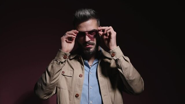 cool fashion model in denim shirt putting sunglasses on, arranging beige jacket and confidently crossing arms in a shadow on pink background in studio