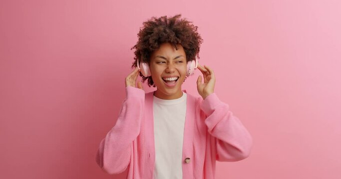 Happy young woman with curly hair dances carefree listens favorite music moves with rhythm feels upbeat enjoys every bit gesticulates with arms dressed casually isolated over pink background
